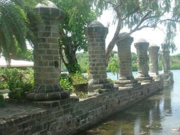 Sideview of famous 6 Nelson's Dockyard pillars, lined up on water front, green trees behind .