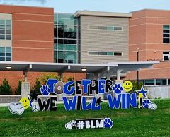 Colorful outdoor sign reads 'Together We Will Win!' with #BLM and smiley faces near a school building.