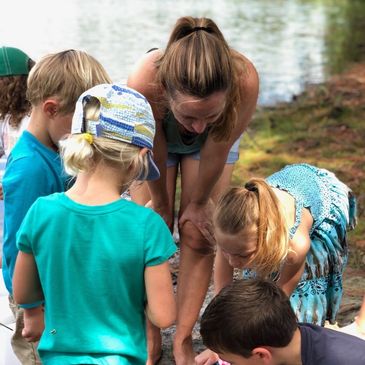 Teacher and students looking down at something on ground with water in background.