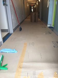 In-process cleaning of long hotel hallway w/floor covered by protective paper. Caulk gun & broom.
