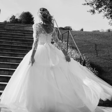 Black and white photo of bride in lace and tulle wedding dress at bottom of steps holding skirt out