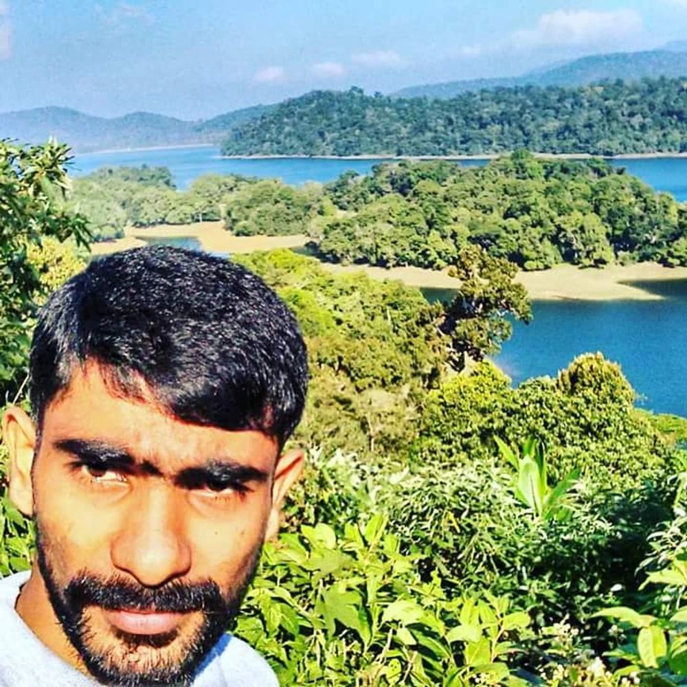 Man taking a selfie with a scenic lake and lush green hills in the background.