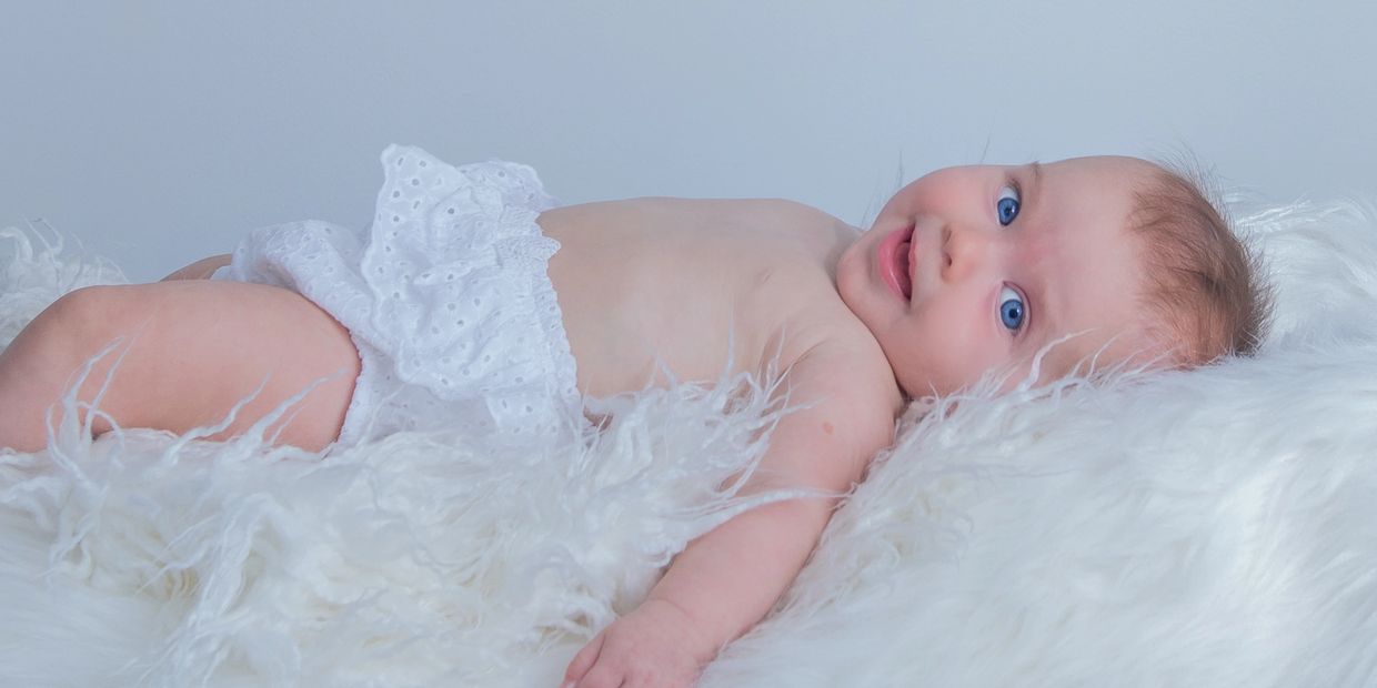 smiling baby girl with bright blue eyes wearing a white diaper cover, lying on a white fluffy rug