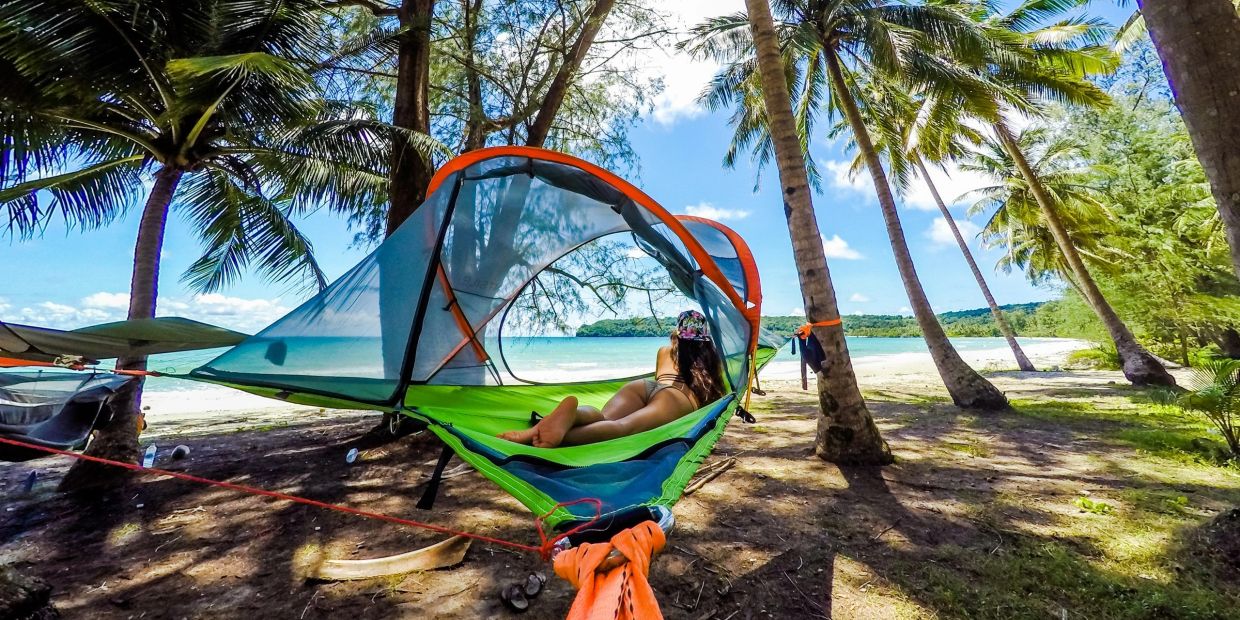 A woman relaxes in a suspended hammock tent by the beach under palm trees.