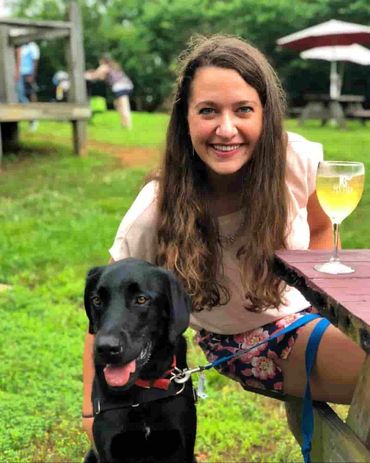 woman at picnic table drinking a glass of white wine with her dog nearby