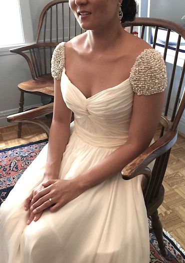 Woman in an elegant white dress with pearl-embellished sleeves sitting on a wooden chair.