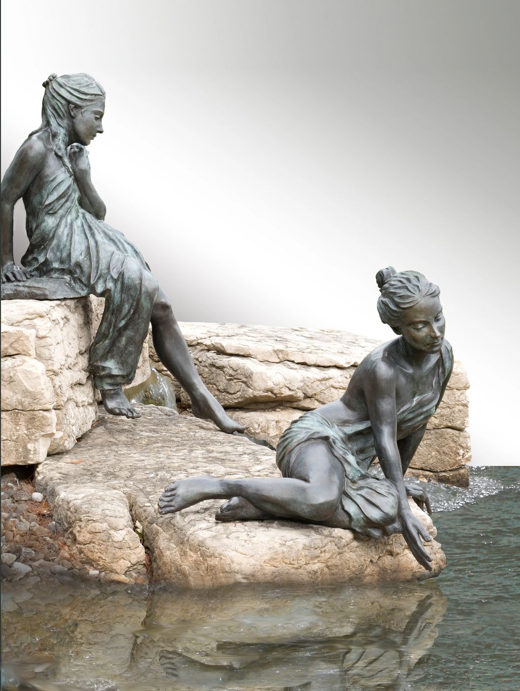 two female figures cast in bronze are seated on rocks while they test the water