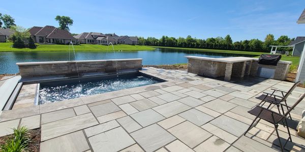 Modern backyard patio with a pool, water fountains, and outdoor kitchen by a lake.