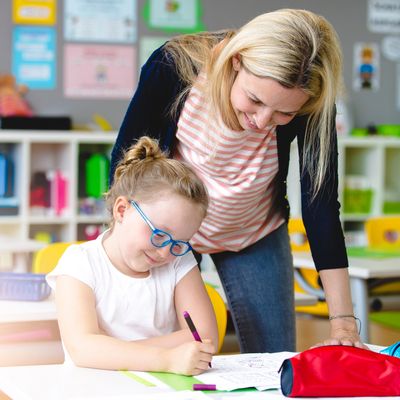 A Speech-language therapist stands next to a girl sitting at a table and writing. They are smiling.