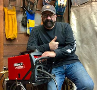 Man in jeans and cap giving thumbs up beside a Lincoln Electric welding machine.