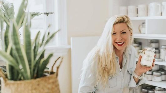 Woman with long blonde hair smiling, holding a candle jar in a bright white room.