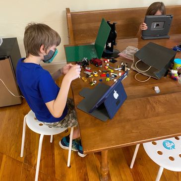 Young students with iPads and legos, making stop-motion videos. The boy has a mask on.