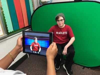 A young student filming in front of a popup green screen. you can see him in an ipad too.