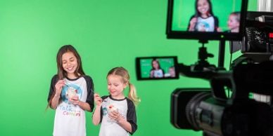Two young students standing in front of a green screen in a studio, eating sundaes.