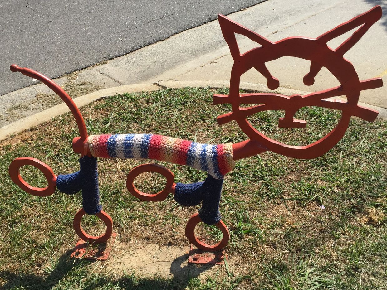 Orange metal sculpture in an abstract shape of a cat with a yarn covering on the body and legs