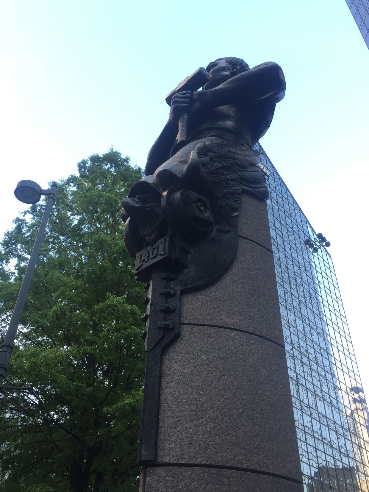 Bronze statue of a strong man clutching a hammer, hands over his chest, kneeling over an eagle.