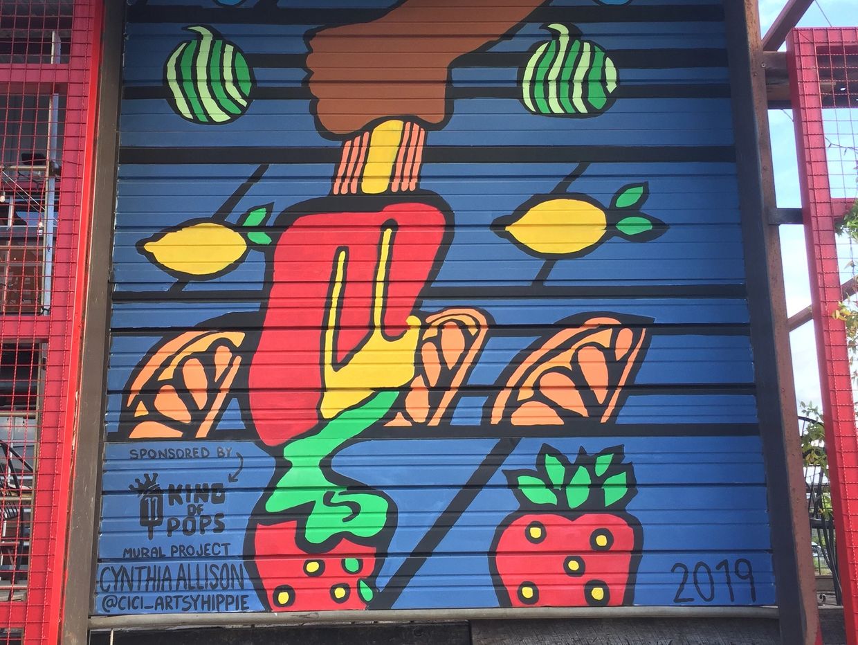 A brown hand holding a red popsicle with strawberries, oranges and lemons around it.