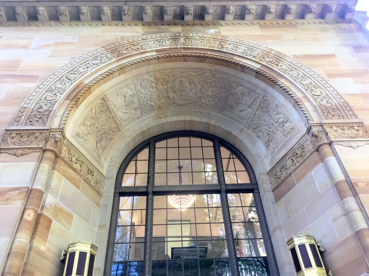 The top of an arched sandstone building entrance intricately carved with classical references.