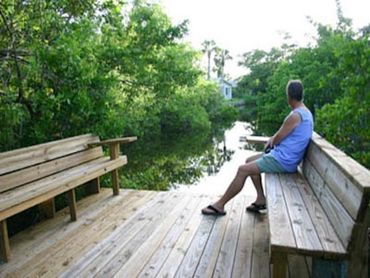 Rich sits on a bench on a wooden dock at a bungalow surrounded by lush greenery and the canal.