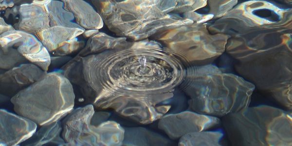 Image of crystal clear lake water with a large water drop and rings emanating out and rocks visible 