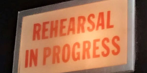 Illuminated sign reading 'REHEARSAL IN PROGRESS' in bold orange letters.