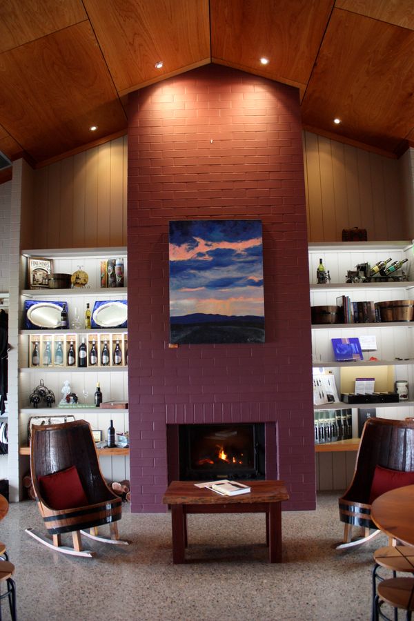 Wine tasting lounge with red brick fireplace, display shelves, barrel chairs, and timber ceiling.