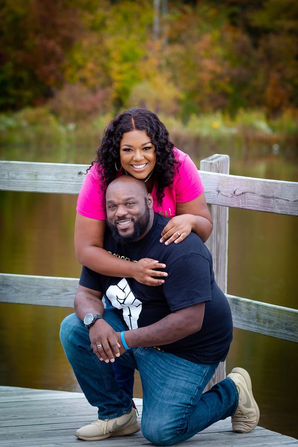 A couple posing on a lakeside deck, with the woman leaning on the kneeling man.