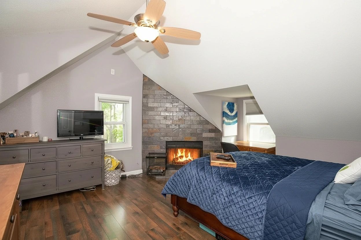 Cozy attic bedroom with vaulted ceiling, stone fireplace with glowing fire, and hardwood floors
