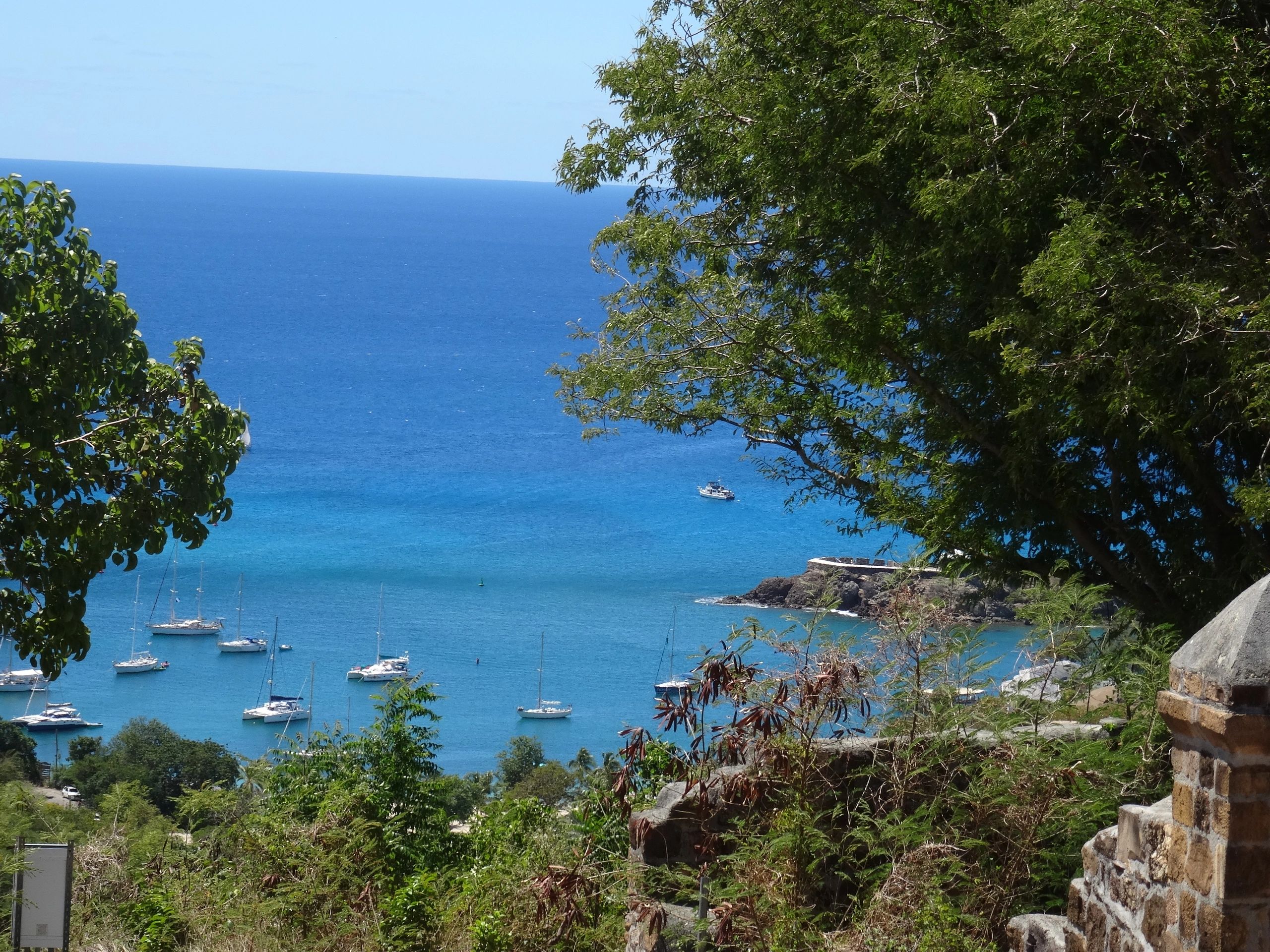 Blick auf Pigeon Point Strand Antigua. Historische Mauer, Bäume im Vordergrund, Meer und Segelboote 