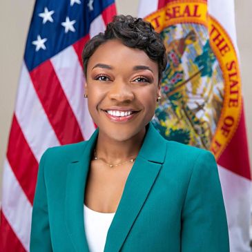Professional woman in teal blazer smiling with American and state flags behind her.