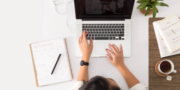 Person typing on a laptop at a tidy desk with a calendar, book, and coffee.
