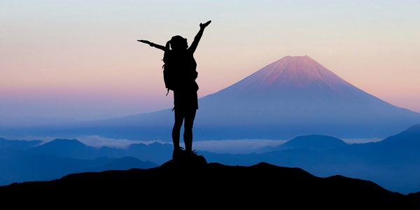 Frau auf einem Berggipfel beim Abendrot, die erfüllt beide Arme in den Himmel streckt