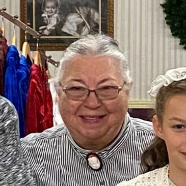 Smiling elderly woman wearing glasses and a striped shirt with a cameo brooch.
