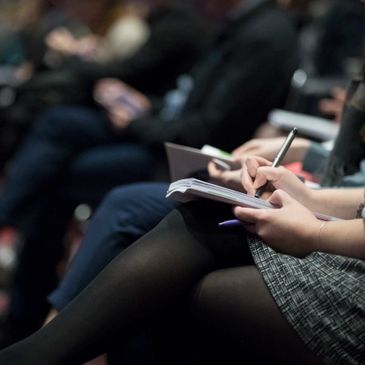 audience members are seated with legs crossed, taking notes. Only legs are shown.