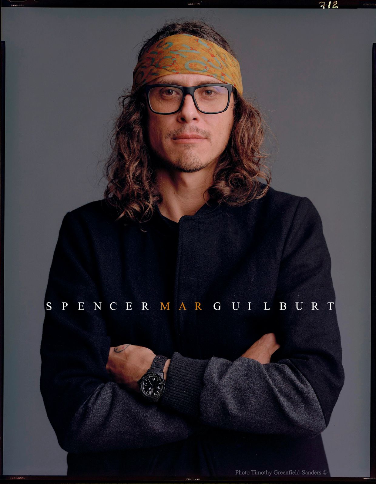 Portrait of a man with long hair, glasses, and a headband, arms crossed.