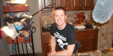 A man smiling in a kitchen with film equipment around him.