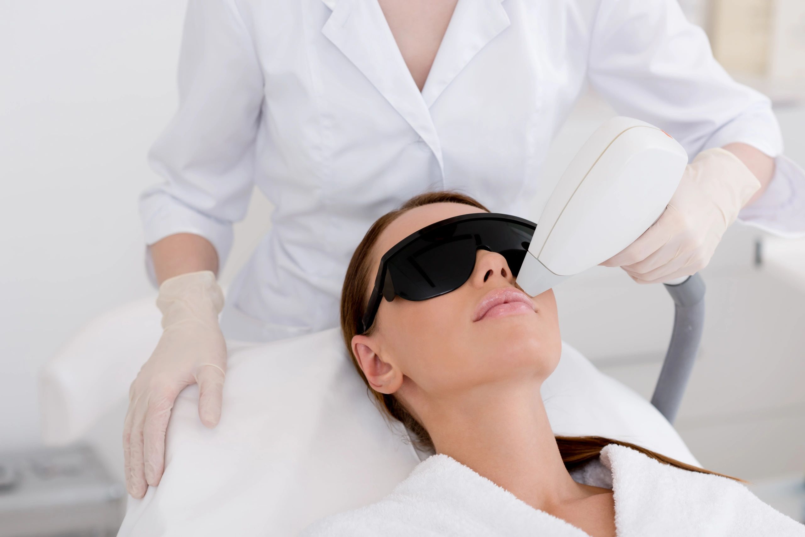 therapist in white overalls performing laser on the lip of a brown haired lady wearing goggles