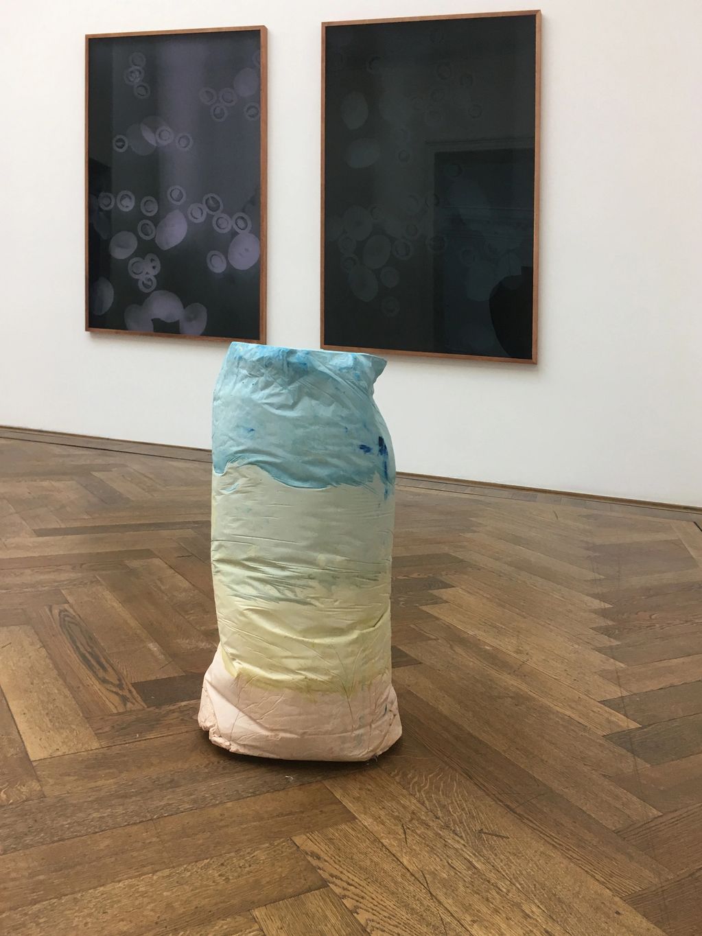 Abstract sculpture with soft colors in front of framed dark artworks in a gallery.