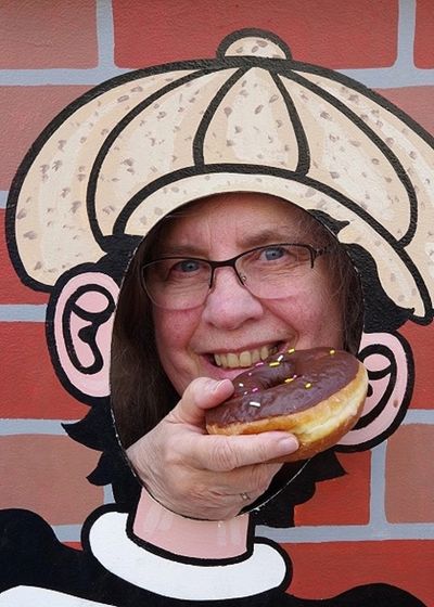 Person posing with face in cartoon cutout, holding a chocolate donut.