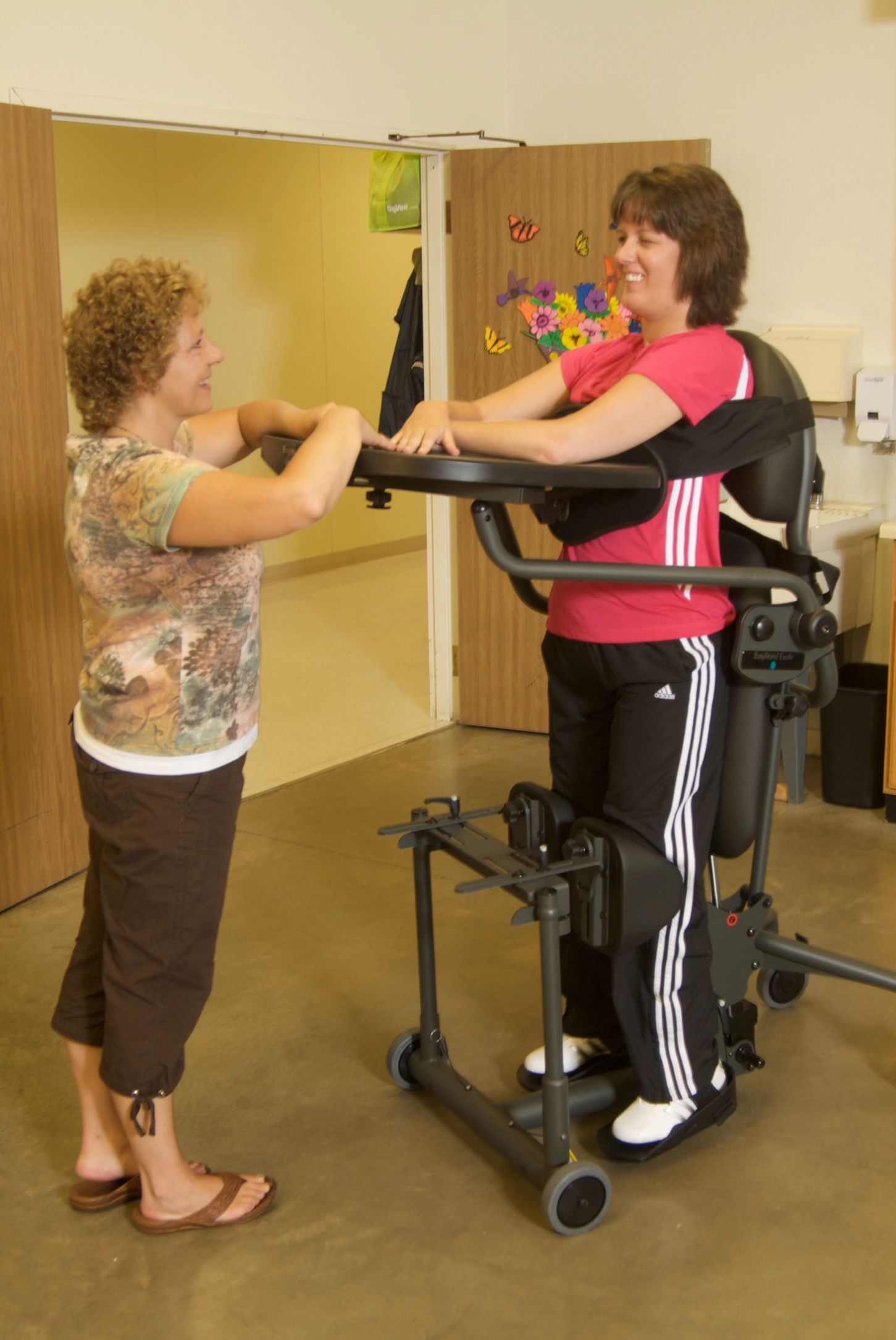 Lady standing in an EasyStand Evolv Shadow Swing-Away standing frame, talking to another lady stanin