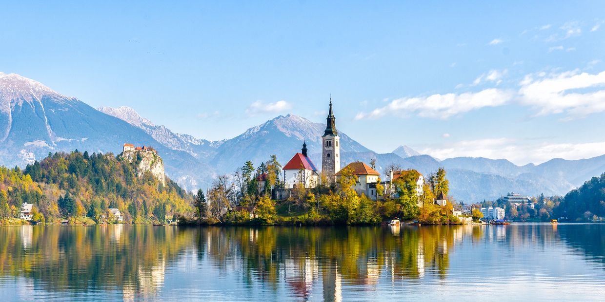 Island with church and buildings reflected in calm lake, surrounded by mountains.