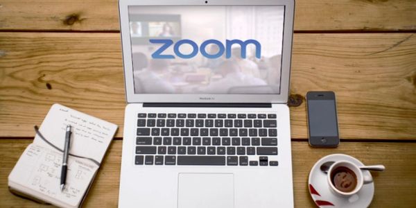 Laptop with Zoom on screen, notebook, phone, and coffee on wooden table.