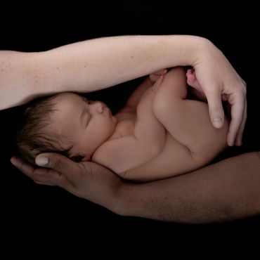 newborn curled up on dark skinned man's arm with light skinned woman's arm above