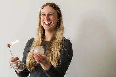 A woman standing in front of a white wall, holding a cup and a fire stick, laughing