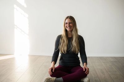 Blond, caucasian woman sitting on wood floor with crossed legs, hands on knees, smiling 