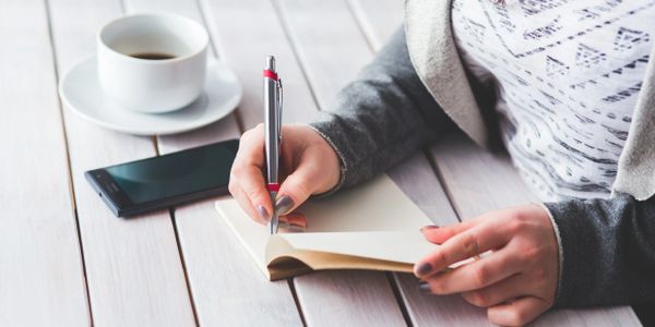 woman in a jacket sitting outside at a table with coffee and writing in a journal