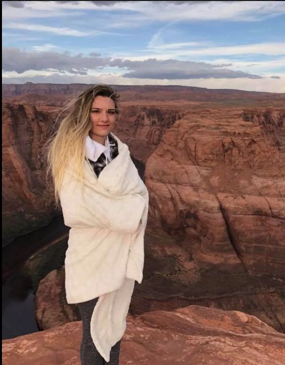 A woman wrapped in a white blanket stands on a rocky cliff overlooking a canyon.