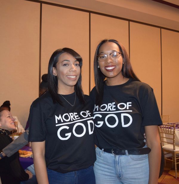 Two women smiling, wearing 'More of God' t-shirts at an event.