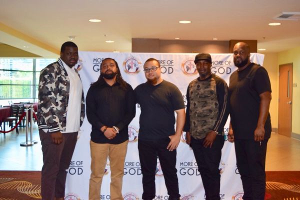Five men posing in front of a 'More of God' backdrop at an indoor event.