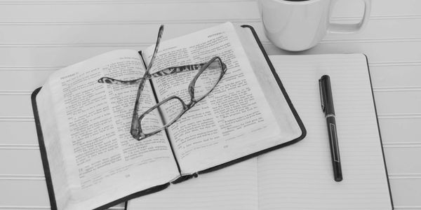Reading glasses resting on an open book beside a notebook, pen, and coffee cup.
